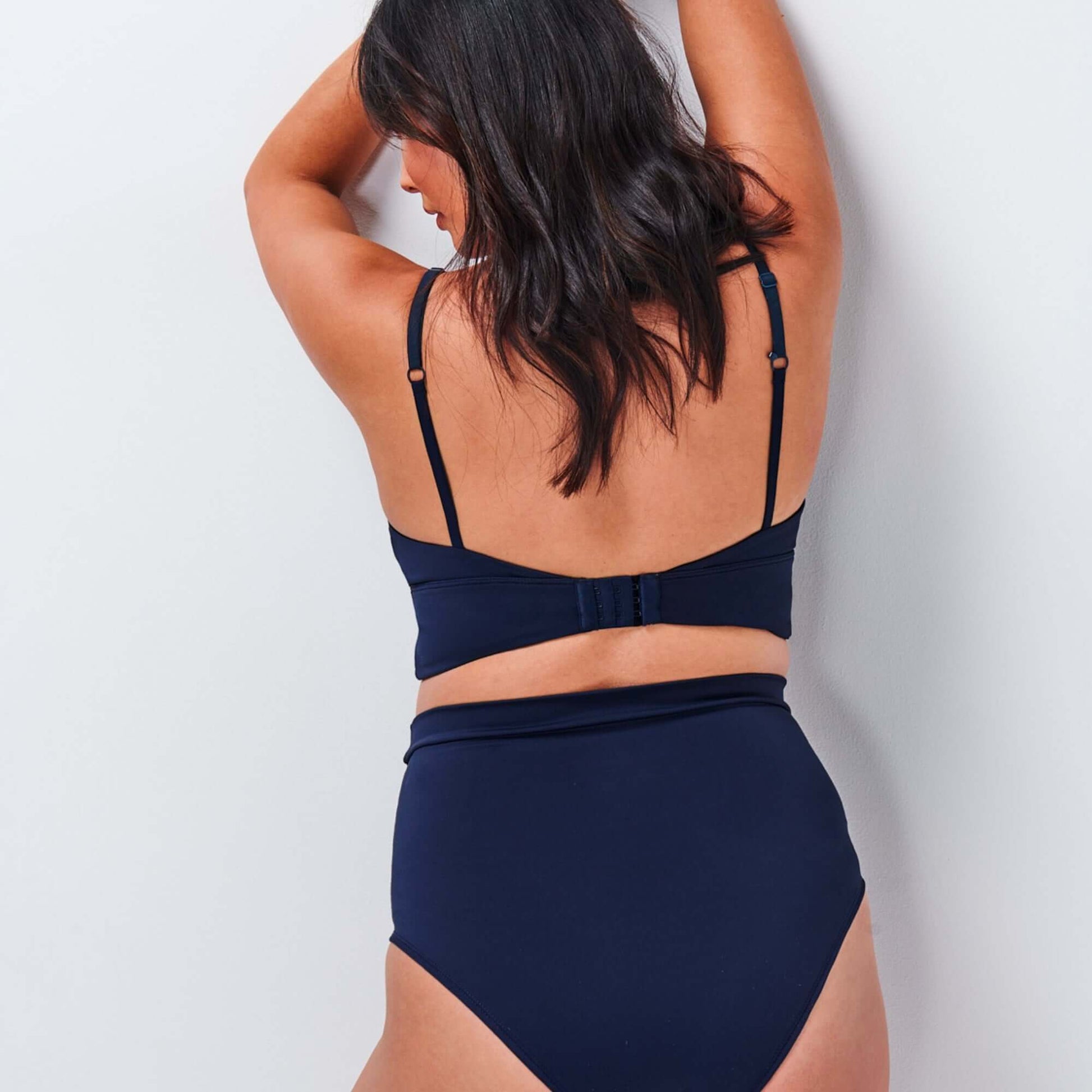 Model faces wall to show the back of the NipCo Maternity Bra and High Waisted Underwear in the colour Navy.
